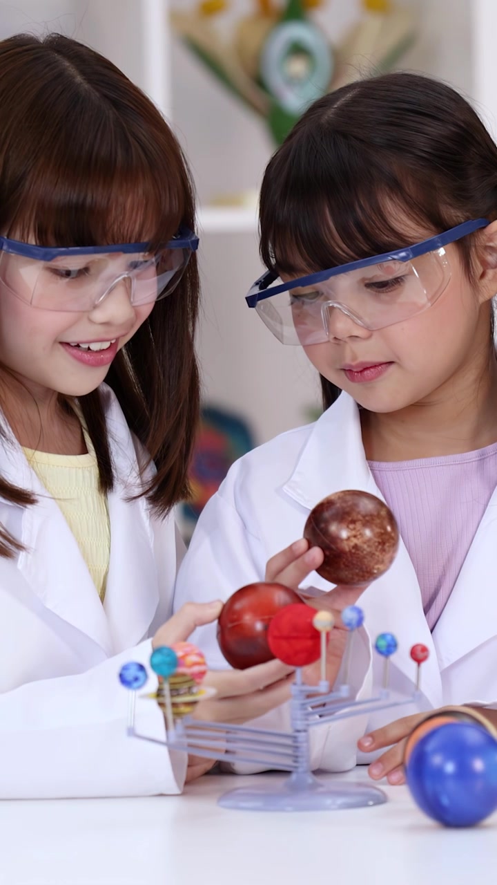 Two young girls wearing safety goggles examine a planetary model in a bright science classroom.