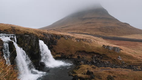 Tiro-De-Seguimiento-Lento-De-La-Cascada-De-Kirkjufellsfoss-Y-Kirkjufell,-Islandia-En-Niebla-Fría