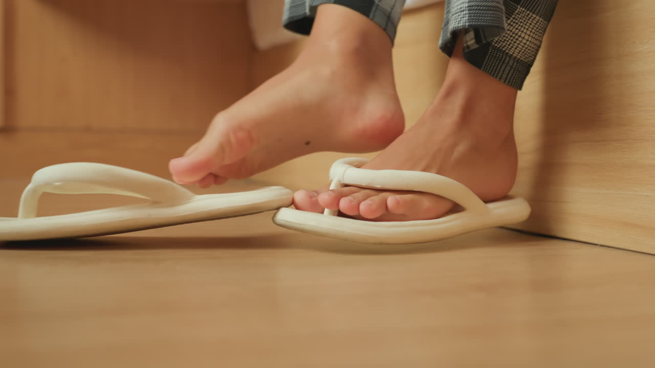 Hotelgast 's nachts op tenen naar pantoffels bij bed, voeten in pyjama bewegen zachtjes over warme houten vloer onder de rand van het bed, close-up van foampantoffels en linnen, intieme schemerige scène die stilte uitstraalt.