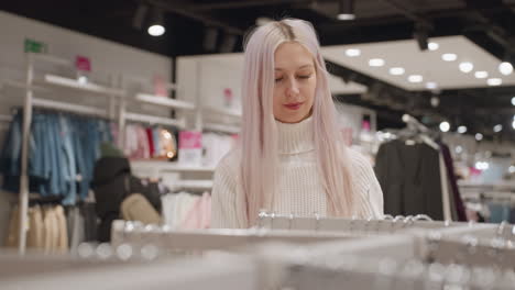 woman in white knitted sweater browsing boutique rack of neutral fleeces under warm lights, pauses to select soft fleece sweater, gently feeling fabric texture as shoppers browse in background