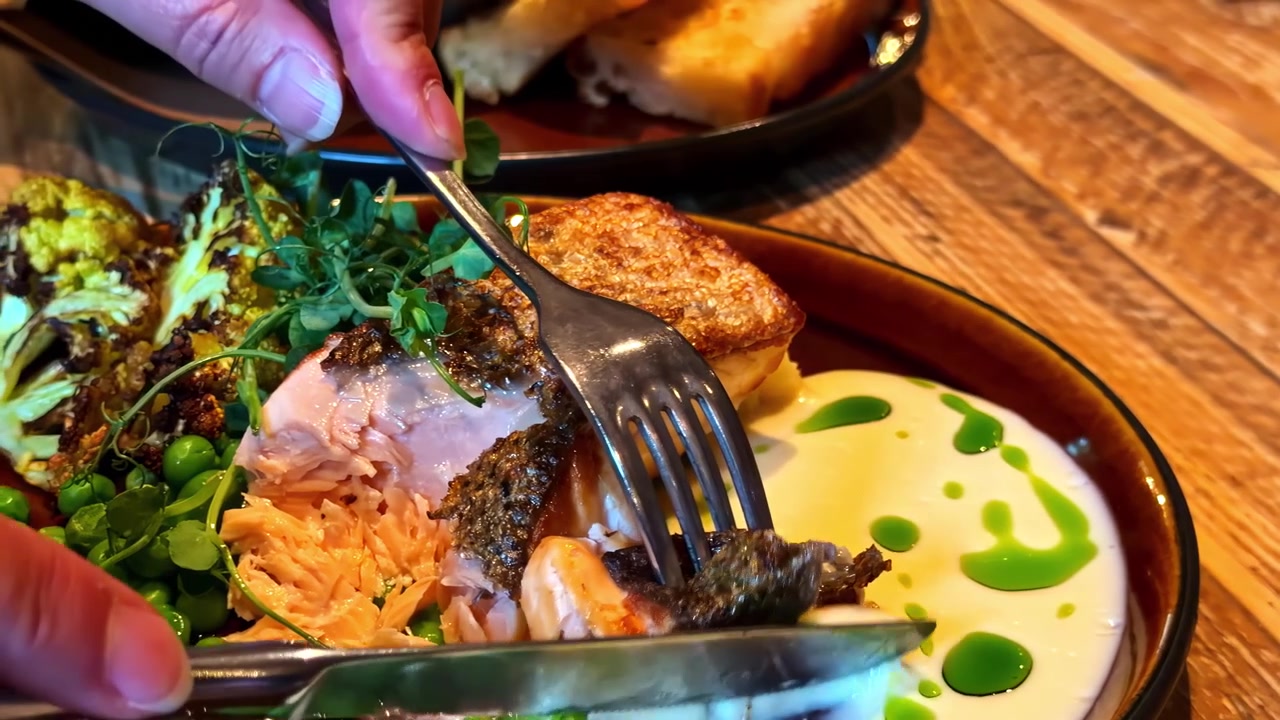 Person cuts into cooked fish fillet with vegetables and sauce at rustic wooden table