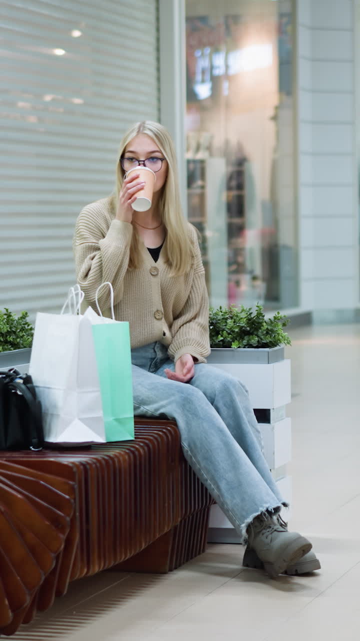 mujer elegante en traje casual sentada en un banco decorativo bebiendo café en un centro comercial moderno, bolsas de compras a su lado, atmósfera relajada con fondo borroso de exhibiciones de tiendas y compradores