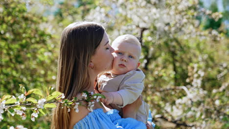 beautiful caucasian woman with long hair wearing blue dress holding a cute blond baby. loving mother kisses her adorable child on the cheek. spring garden at backdrop.