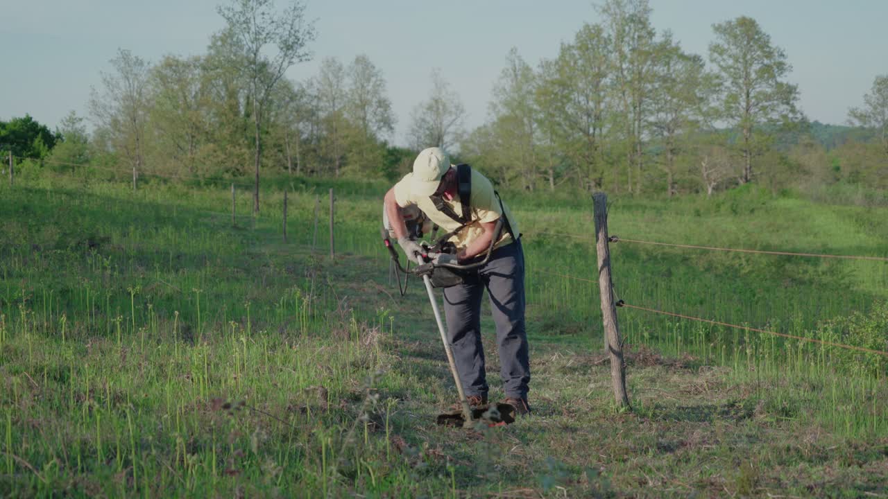 Worker clears weeds with a brush cutter beside fence in sunny countryside field