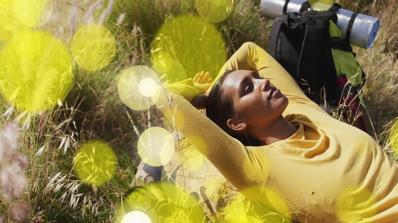 Panoramica della telecamera su una scena di benessere in un prato, mentre una donna sdraiata su un tappetino respira, con riflessi di luce fluttuanti