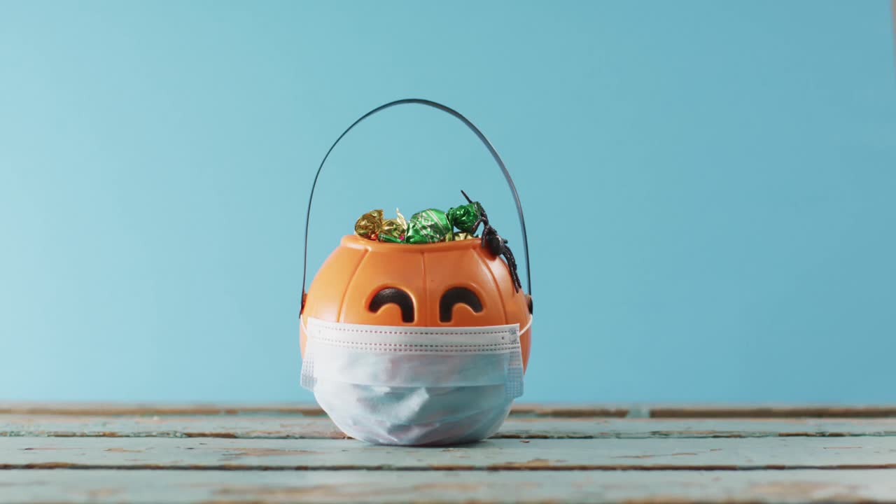 máscara de cara sobre un cubo en forma de calabaza lleno de dulces en una superficie de madera contra un fondo azul