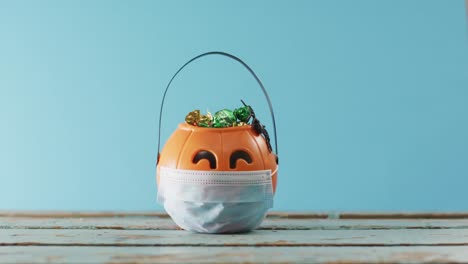 Face-mask-over-pumpkin-shaped-bucket-full-of-candies-on-wooden-surface-against-blue-background