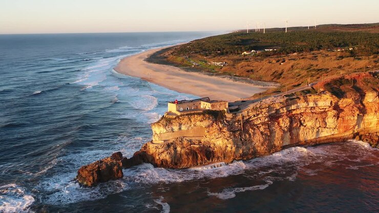 Faro a Nazaré, Portogallo, vicino a Praia do Norte durante il tramonto, ripresa aerea circolare in allontanamento.