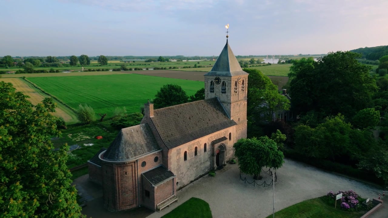 Luftaufnahme der historischen Alten Kirche in Oosterbeek, Niederlande, gebadet im Licht der goldenen Stunde. Umgeben von üppig grünen Feldern, verstreuten Bäumen und der mäandernden Flusslandschaft, die die...