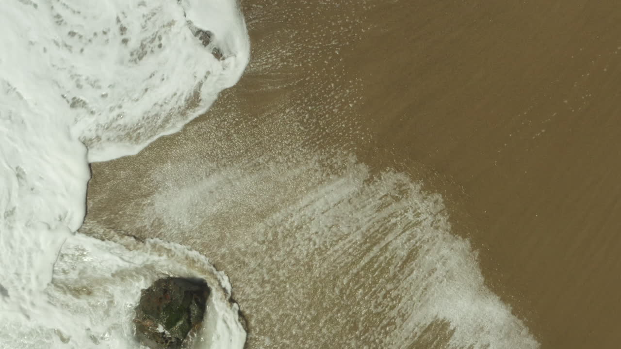 Ripresa aerea ascendente di onde spumeggianti che si infrangono sulla spiaggia sabbiosa.