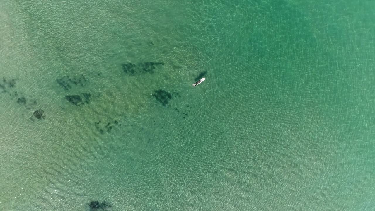 드론이 회전하면서 상승하면서 파도가 없는 맑은 물에서 보드에 누워 있는 서퍼의 오버헤드 뷰