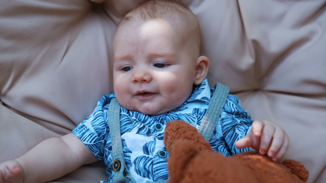 Adorable Caucasian infant in blue shirt lies on the soft backdrop. Fluffy brown teddy is near the kid. Close up.
