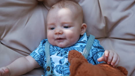 adorable caucasian infant in blue shirt lies on the soft backdrop. fluffy brown teddy is near the kid. close up.