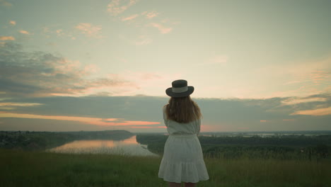 back view of an adult woman in a white dress and hat standing alone in a serene grassy field, gazing out over a tranquil lake during sunset under a soft, pastel-colored sky