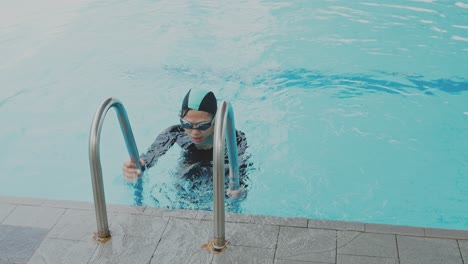 Young-Asian-Woman-In-Black-Swimsuit-And-Swim-Goggles-Getting-Out-Of-Pool-Using-Steps