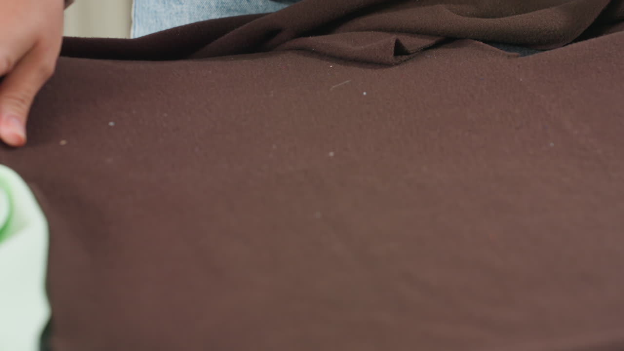 Dress designer spreads material neatly across worktable adjusting thick dark brown fabric carefully while positioning white cut out pattern for accurate garment preparation inside sewing studio