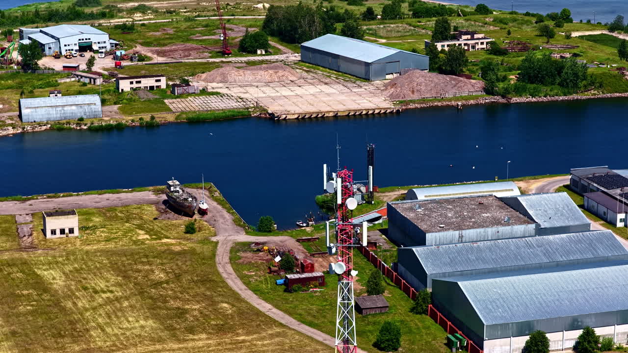 Aerial shot flies past a telecommunications tower to reveal the industrial harbor of Mērsrags Port, Latvia, with its timber storage yards, warehouses, and docked ships