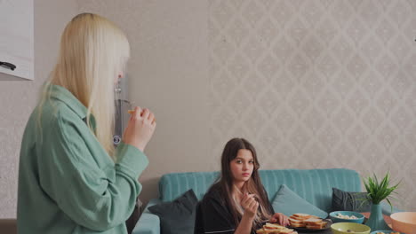 two women enjoy breakfast together in cozy kitchen setting, one sitting on sofa with plate of bread and cheese while other stands nearby holding sandwich as she eat it