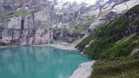 Aerial-descend-drone-shot-above-shore-line-between-pine-trees-and-famous-turquoise-azure-glacier-lake-Oeschinensee-surrounded-by-Bluemlisalp-giant-mountains-in-Kandersteg,-Switzerland