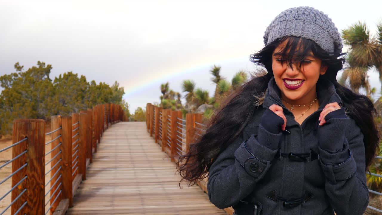 una joven feliz y hermosa caminando por el desierto con un arco iris en el cielo nublado durante una tormenta de lluvia a cámara lenta