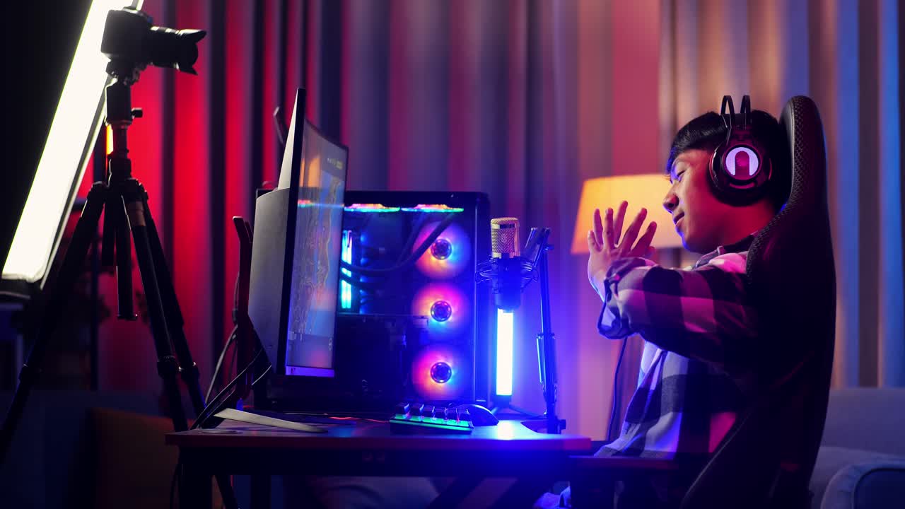 vista lateral de un streamer asiático que se estira mientras juega un juego a través de la red en una computadora personal. videojuego de transmisión en vivo, escritorio iluminado por luz de tira led rgb