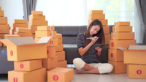 a young woman sits among shipping boxes doing inventory and recording them on a clipboard