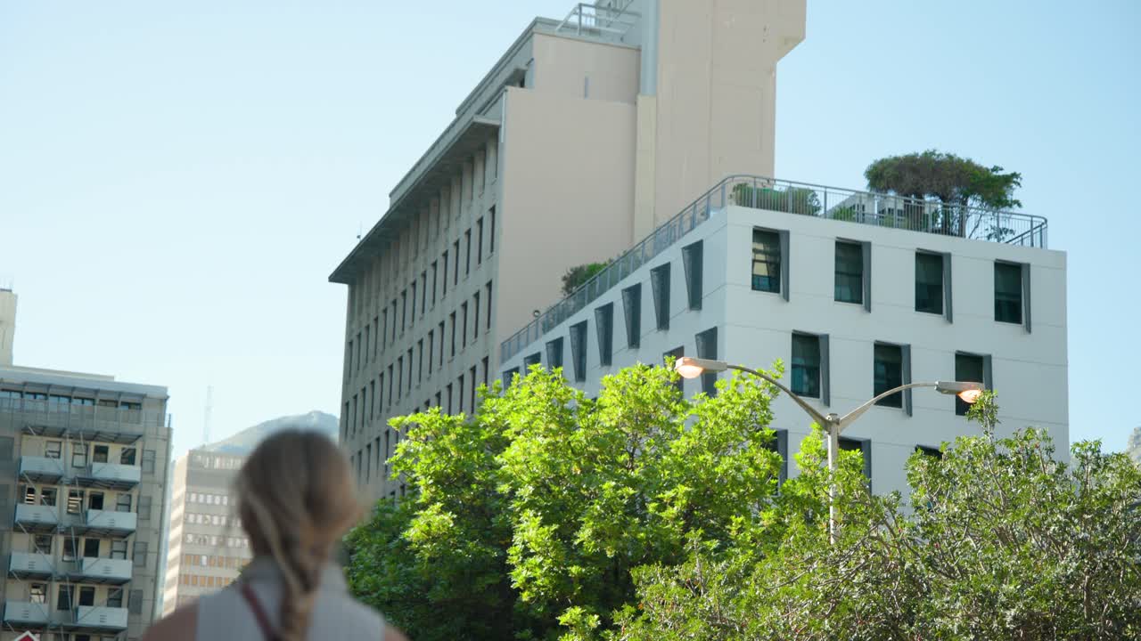 Edifício bege e árvores margeiam a rua, enquanto duas pessoas que entram no enquadramento caminham com café.