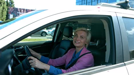 beautiful blonde hair woman wearing pink long sleeve driving the car while smiling on the camera - close up shot