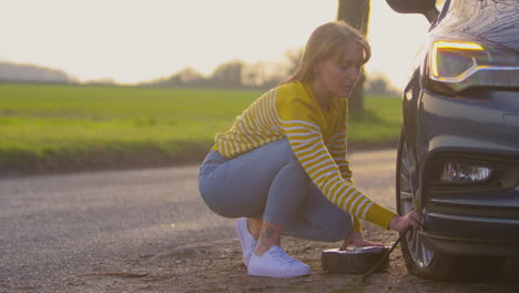 woman inflating car tyre with electric pump on country road