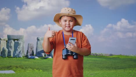 asian boy with a hat and binoculars smiling and pointing to smartphone while traveling in stonehenge. boy researcher examines something, travel tourism adventure concept