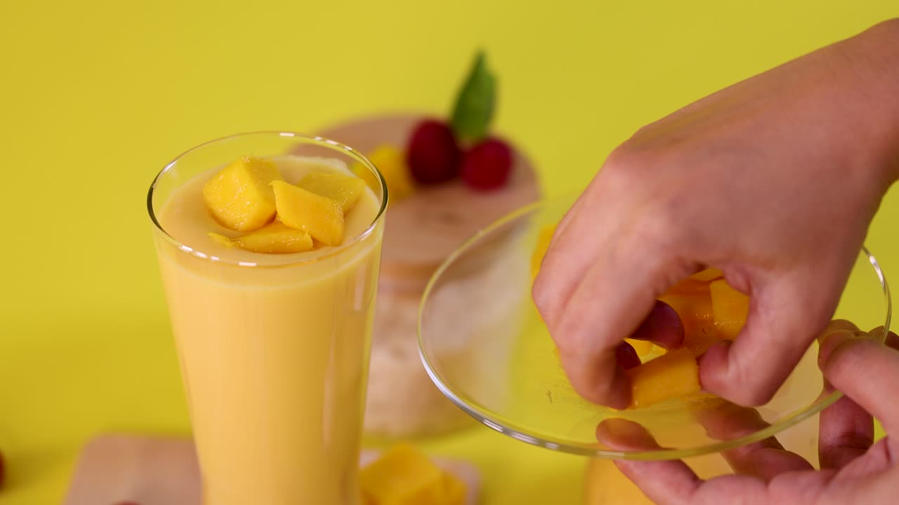 Hand places mango cubes onto smoothie in glass, bright yellow background, overhead close-up shot