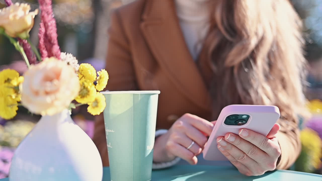 Woman in brown coat scrolling on her phone near a cup of tea at a terrace