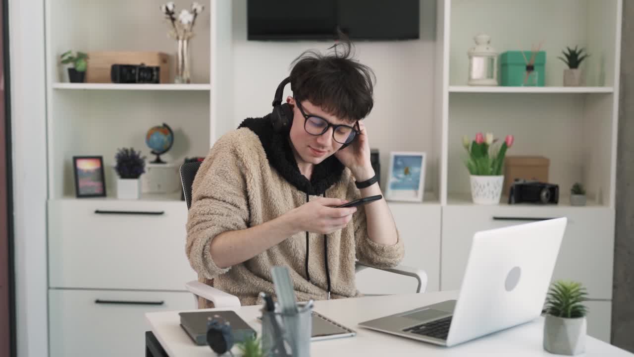 The young man is working on a laptop in the office. He is multitasking, talking on the phone, typing on the laptop, and taking notes on the tablet.