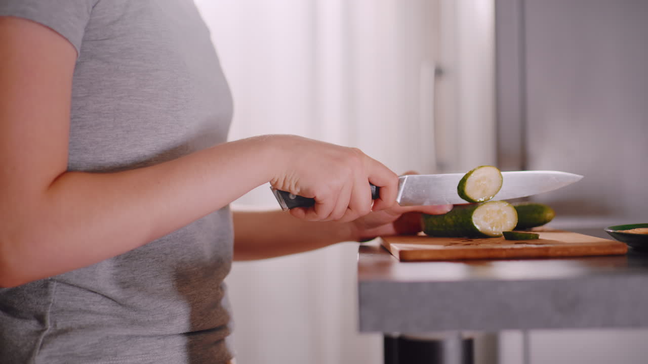 Housewife wearing grey shirt slices cucumber on cutting board in kitchen with fridge in background, light shining softly through curtains, showing calm domestic routine