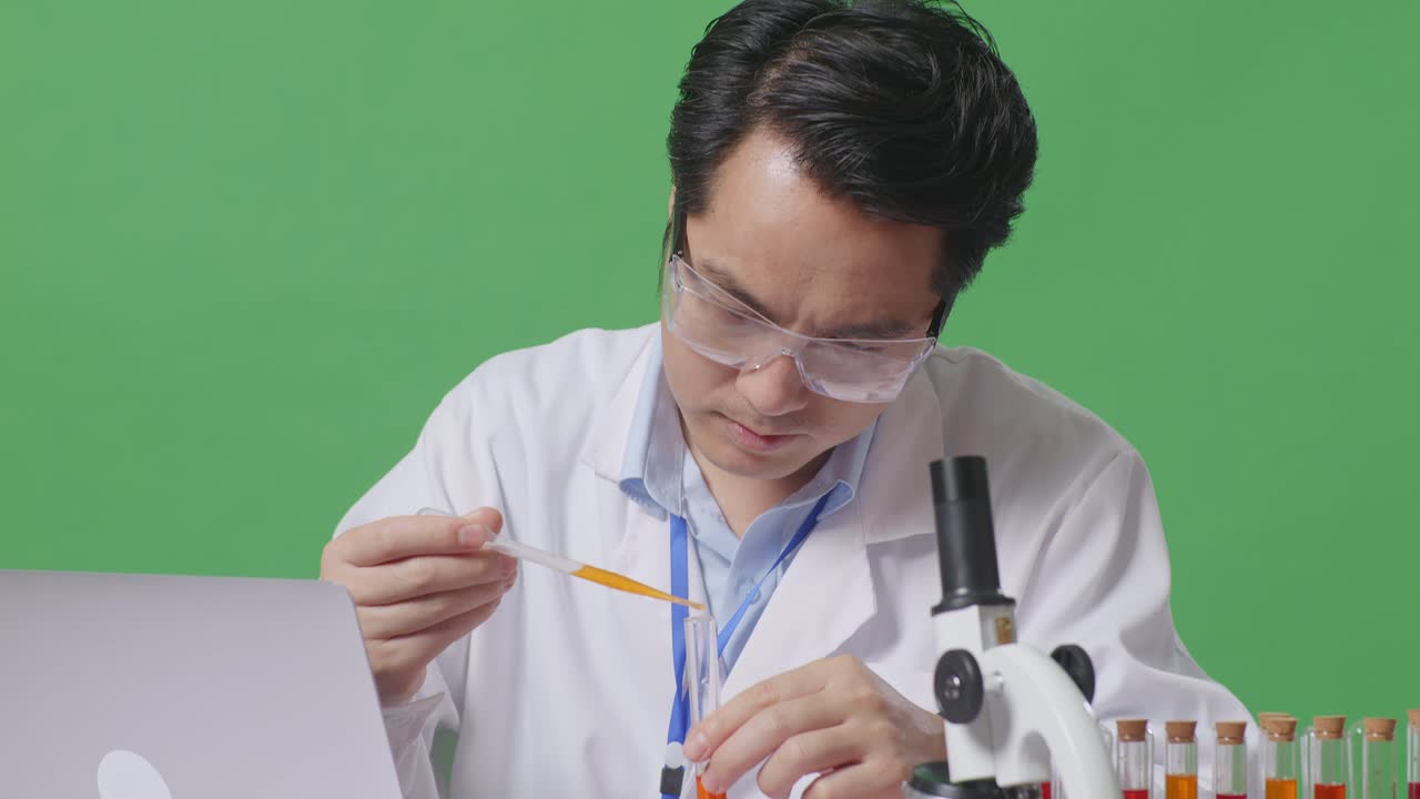 Close Up Of Asian Man Scientist Making Experiment With Test Tube And Typing On A Laptop While Working On The Table With Microscope In The Green Screen Background Laboratory