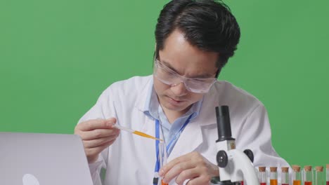 close up of asian man scientist making experiment with test tube and typing on a laptop while working on the table with microscope in the green screen background laboratory