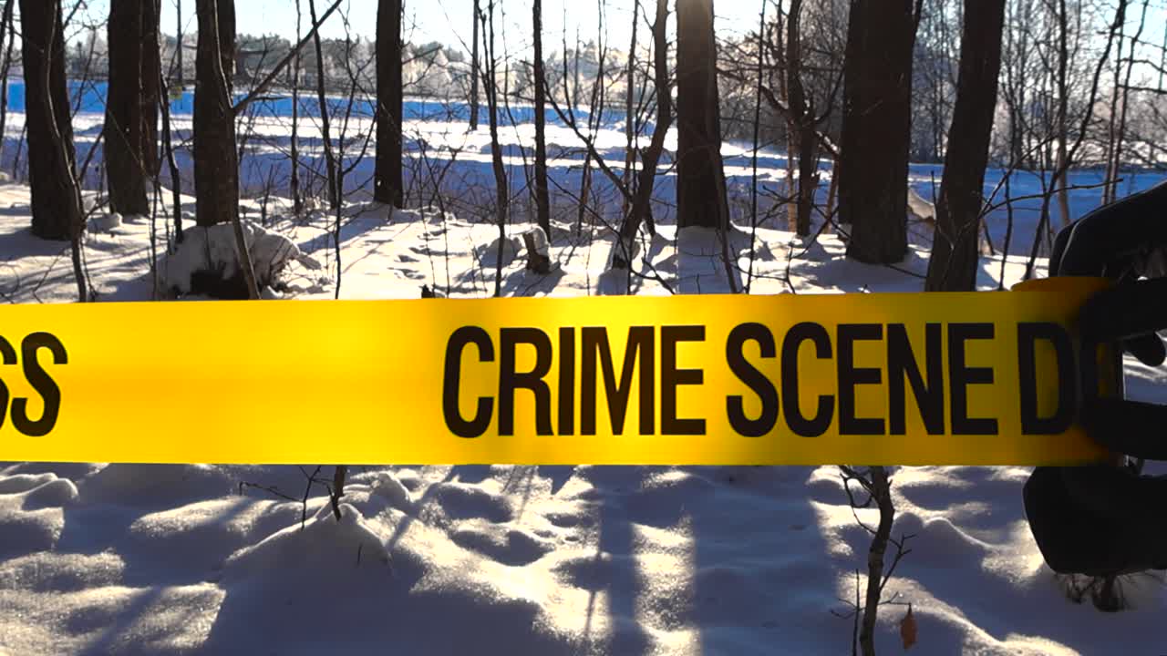 Gloved investigator hand unrolls caution tape through sunlit snowy woodland, marking a restricted forensic area