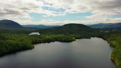 Aerial-Drone-Footage-of-Loch-an-Eilein-in-the-Cairngorms-National-Park-Scotland-Revealing-a-Native-Scots-Pine-Forest-and-Mountains-Behind-on-Blue-Sky-and-Cloudy-Day