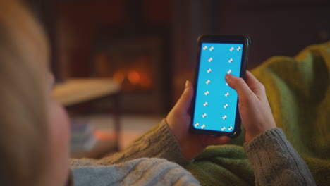 mujer en casa acostada en el sofá en la sala de estar con fuego acogedor usando teléfono móvil de pantalla azul