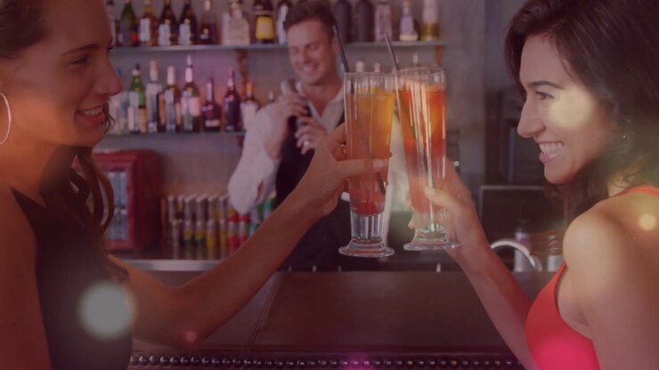 Due donne che alzano i bicchieri, brindando in un bar, con luci bokeh che illuminano i bicchieri mentre il barista solleva lo shaker