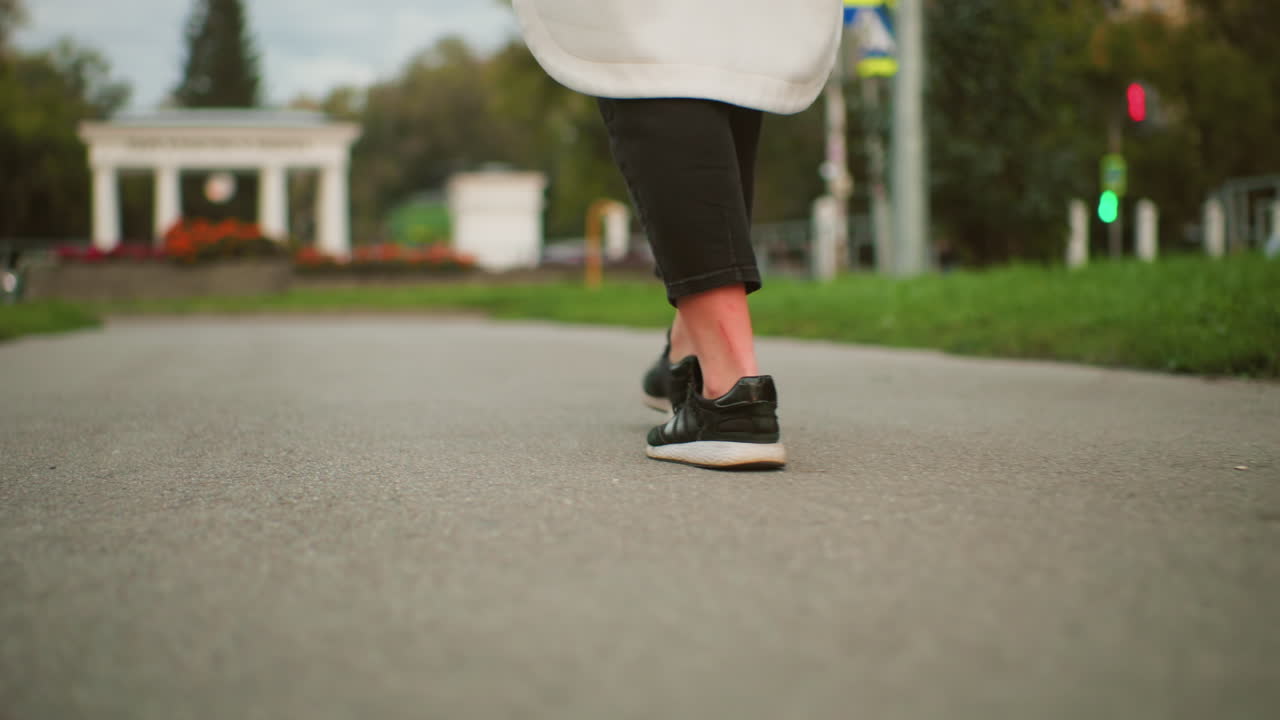 Lower body of lady wearing long coat and sneakers walking while twirling closed umbrella, blurred city background with green grass, traffic light, distant moving cars in urban setting