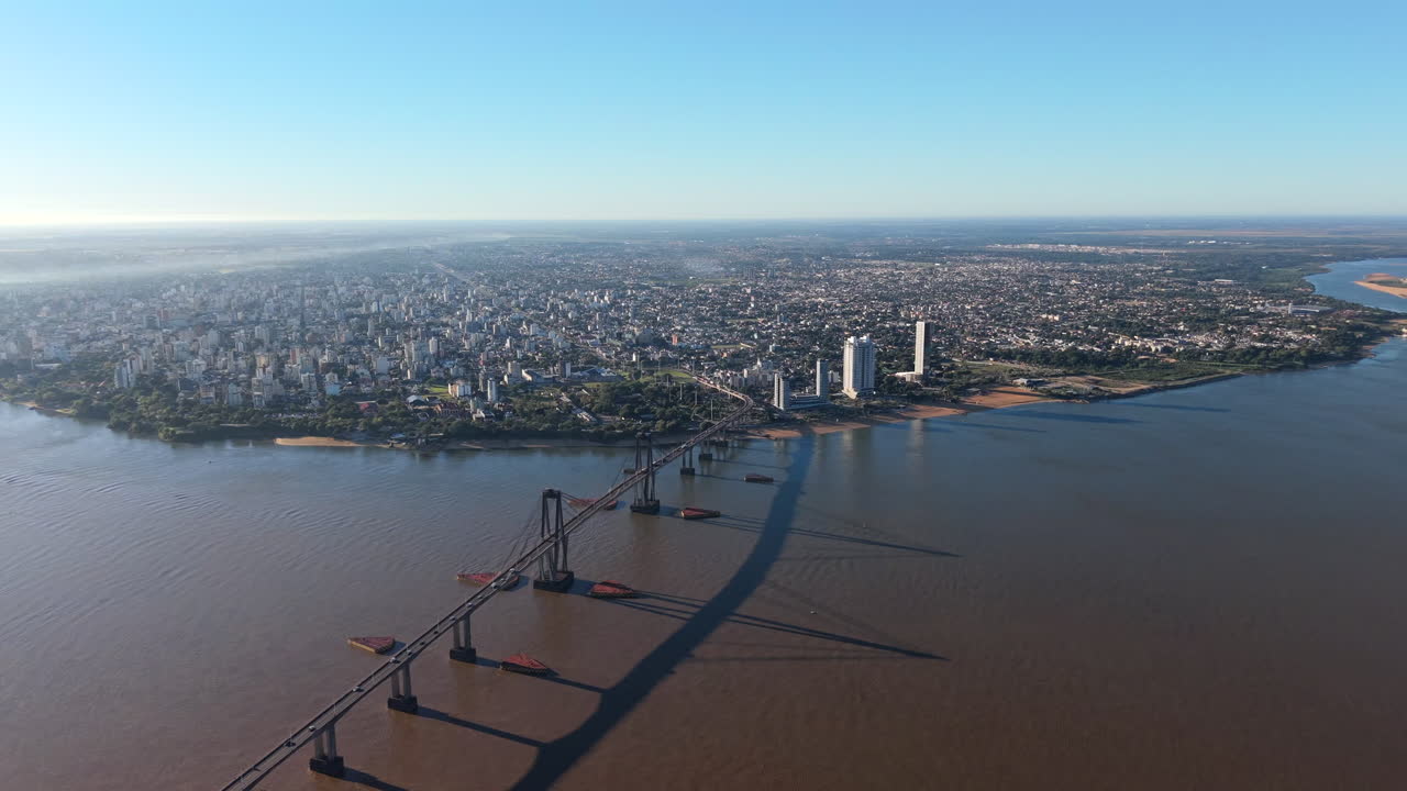 Drone ad altissima quota in discesa in avanti, che cattura l'intera scena: ponte, fiume, città e ombre mattutine che si fondono nel lontano orizzonte