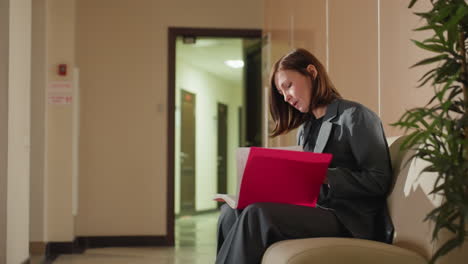businesswoman in gray suit sitting on bench in office corridor deeply focused on reviewing paperwork inside bright pink folder during quiet moment of concentration in professional environment indoors