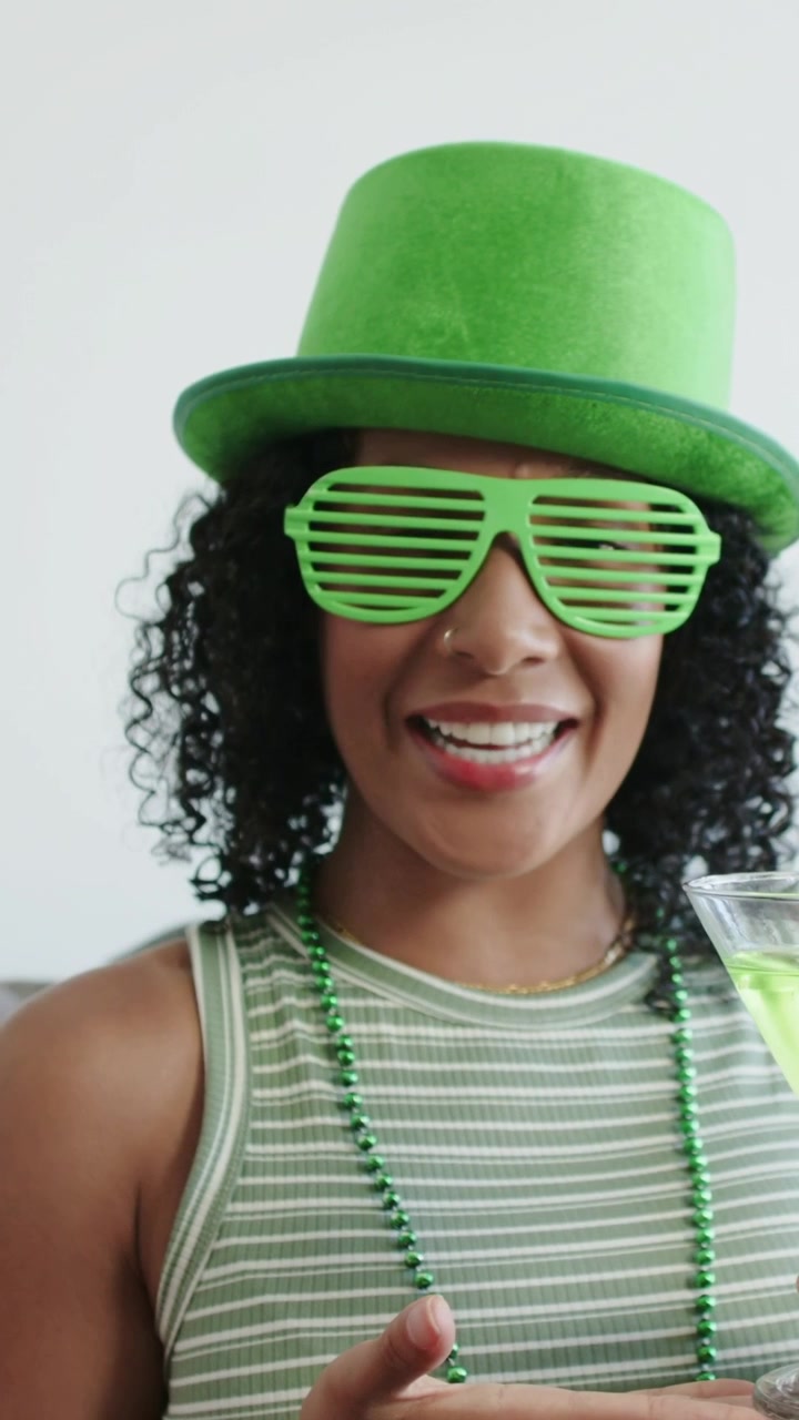 Vertical video: Smiling woman with hat and beads showing green drink on couch, camera pulling back