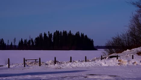 Congelado-Sobre-El-Lago-Astotin-Dentro-Del-Parque-Provincial-De-La-Isla-De-Los-Elk-Con-La-Isla-De-La-Frambuesa-En-El-Horizonte-Mientras-Los-Excursionistas-De-Invierno,-Padre-E-Hijo,-Caminan-Justo-Al-Lado-Del-área-De-Estacionamiento-Público-Con-Bancos-Que-Cercan-Los-Postes-2-2