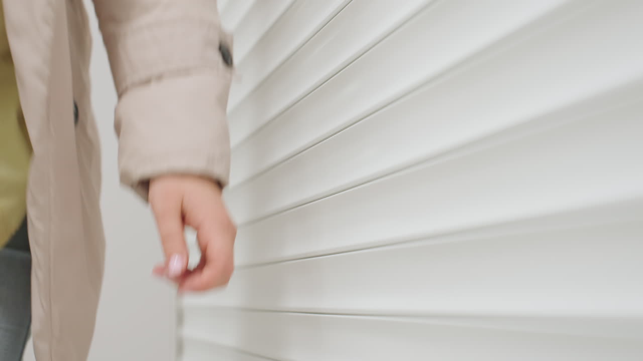 Female manager with light brown hair in jacket rolling down steel shutter to close laundry room, indoor corridor setting, dim background, morning or evening routine in service facility