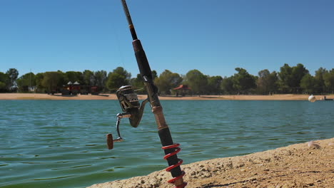 fishing rod anchored in rocky shore by blue lake, slow motion pan