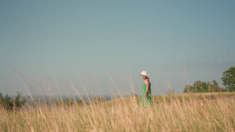 woman in green gown and white sunhat walks peacefully through dry golden grass under soft sunlight, serene side view against expansive sky and distant horizon