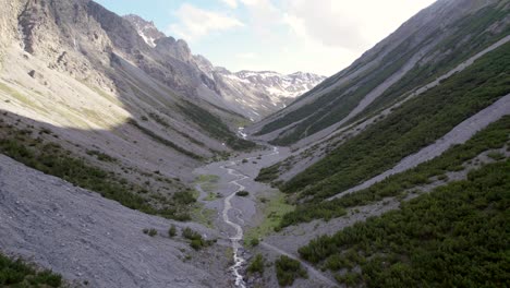 aerial drone footage flying down and through a dramatic glacial valley surrounded by a steep mountains and pine trees with patches of snow and an alpine river in switzerland