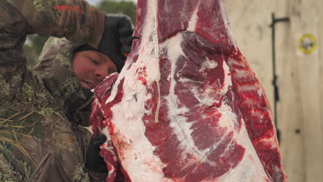 a hunter dressed in a warm camouflage coat is seen cutting away at a fresh hunk of raw meat as it hangs outdoors, close up shot